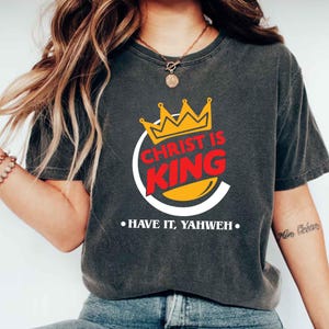 May include: Dark gray t-shirt with a graphic that reads "CHRIST IS KING" in red and yellow, with a crown above the text. The shirt also has the text "• HAVE IT, YAHWEH •" below the graphic.