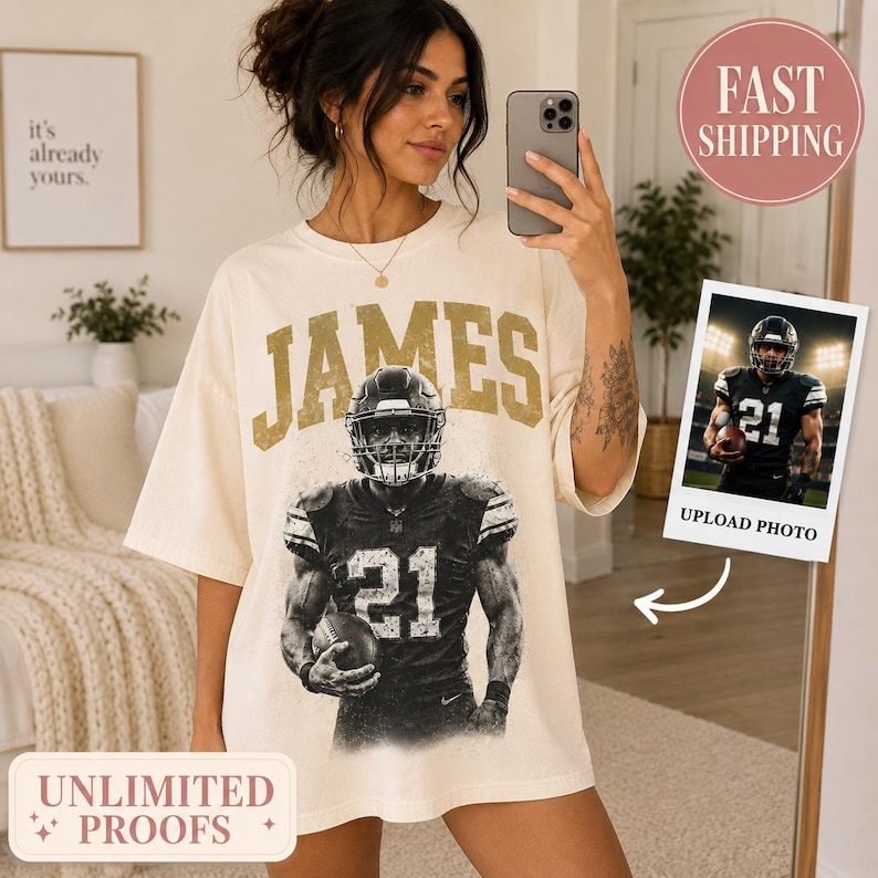 May include: An off-white short-sleeve t-shirt featuring a large graphic print of a football player wearing a jersey with the number 21 and the name JAMES in gold block letters above the player. The shirt has a crew neckline and a loose fit.