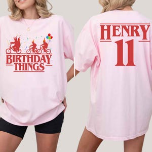 May include: A light pink t-shirt with a graphic on the front that says "BIRTHDAY THINGS" and features three figures riding bicycles. The back of the shirt has the name "HENRY" and the number "11" in red.