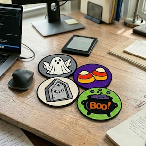 May include: Four circular embroidered patches featuring Halloween designs: a white ghost on a grey background, two orange and yellow candy corns on purple, a grey tombstone with R.I.P. text on cream, and a black cauldron with the word Boo! on green.