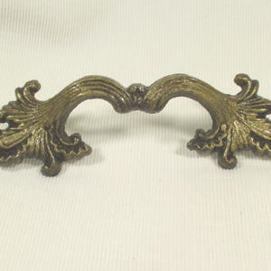 May include: A brass-toned metal drawer pull featuring an ornate, curved design with detailed leaf-like patterns on both ends. The handle has a central arch and two mounting points, showcasing a weathered, antique-style finish with dark highlights.