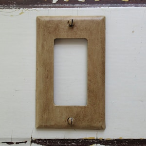 May include: A light brown, rectangular wall plate with a square opening in the center. The plate has two small, black screws on the top and bottom.