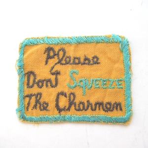 May include: A rectangular patch with a mustard yellow background and teal stitching around the edges. The text "Please Don't Squeeze The Charmin" is embroidered in dark brown and teal thread.