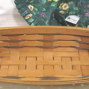 May include: A rectangular woven basket featuring light tan wood splints with dark grey accent strips. The basket includes two brown leather handles attached to the sides. A dark green fabric liner with a yellow and blue floral pattern is visible.