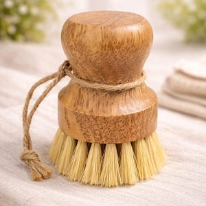 May include: A round wooden dish brush with a light brown wood grain handle and a thick, circular base of stiff, light-colored bristles. A thin, natural-colored jute twine cord is wrapped around the neck of the handle and hangs off the side.