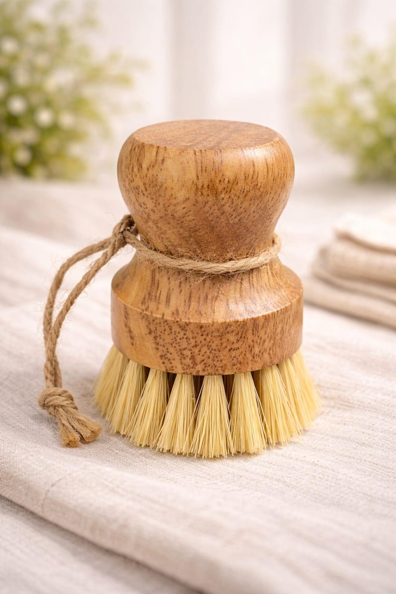 May include: A round wooden dish brush with a light brown wood grain handle and a thick, circular base of stiff, light-colored bristles. A thin, natural-colored jute twine cord is wrapped around the neck of the handle and hangs off the side.