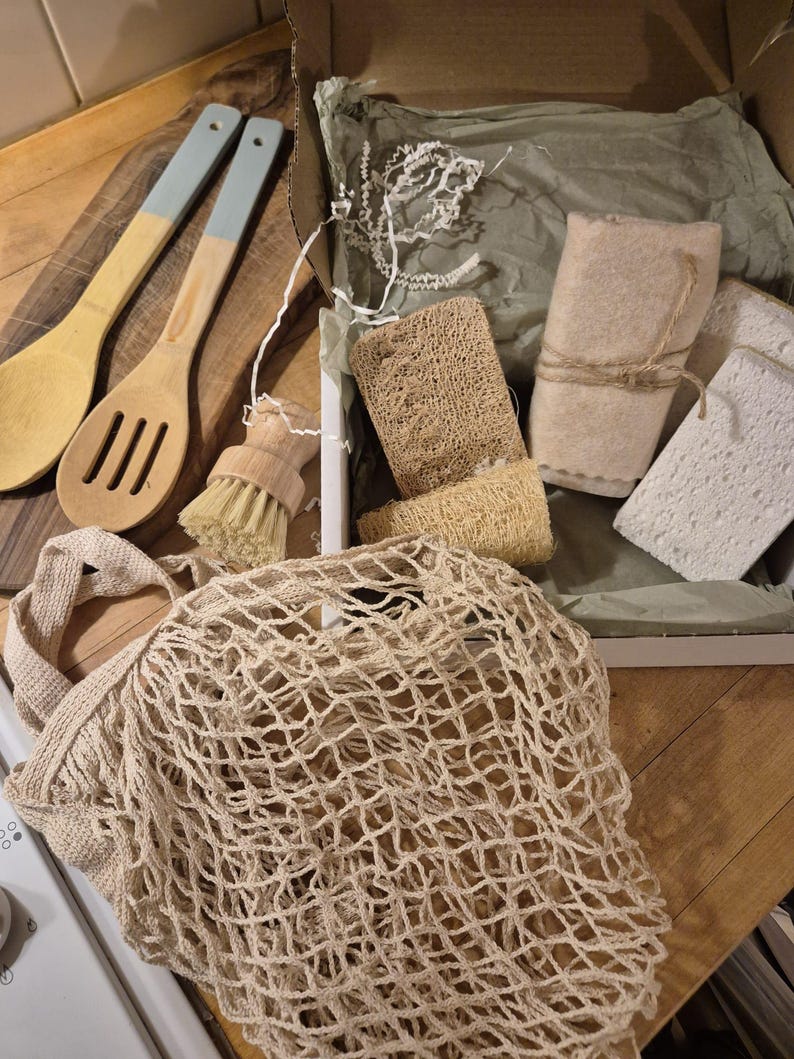 May include: A kitchen set containing a beige cotton mesh market bag, two wooden spoons with light blue handles, a small round wooden scrubbing brush, two textured loofah sponges, and two white rectangular sponges inside a cardboard box.