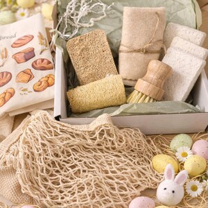 May include: A gift box containing a wooden scrub brush, a beige felt cloth, two loofah sponges, and three white rectangular sponges. Beside the box is a beige mesh tote bag, a cloth with bread illustrations, and several pastel-colored speckled eggs.