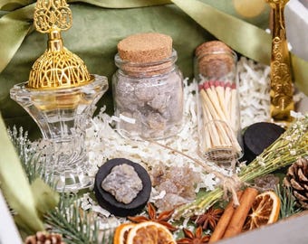 May include: A gift box containing a gold-toned metal incense burner, a glass jar with a cork lid filled with resin, a glass vial of matches, a gold-toned metal spoon, pinecones, dried orange slices, cinnamon sticks, and star anise.