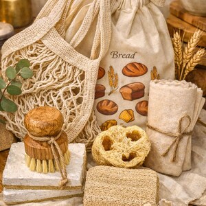 May include: A beige drawstring bread bag with a printed bread illustration and the word Bread, a beige mesh produce bag, a wooden dish brush, two loofah sponges, and a rolled beige cloth tied with twine, all arranged on a light fabric surface.