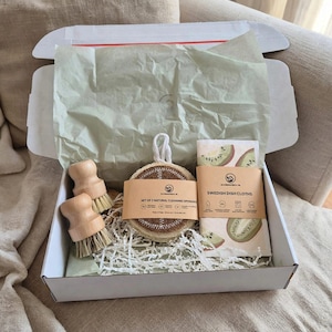 May include: A white cardboard box containing two round wooden scrub brushes, a set of three round natural cleaning sponges with a paper band, and a pack of Swedish dish cloths with a kiwi fruit pattern, all nestled on white paper shred.