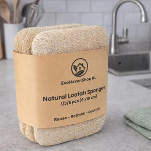 May include: A stack of rectangular natural loofah sponges wrapped in a tan paper band. The band features the text EcoHavenShop NL, Natural Loofah Sponges, 1/3/5 pcs (6x10 cm), and Reuse - Rethink - Reduce, along with a circular house and leaf logo.
