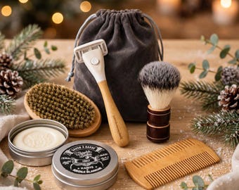 May include: A shaving kit featuring a razor with a light wood handle, a shaving brush with dark bristles, a wooden comb, a round tin of Savon à Barbe shaving soap, and a small oval brush, all arranged in front of a grey drawstring pouch.