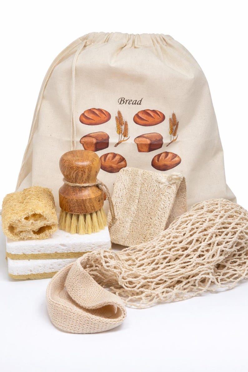 May include: A beige drawstring bread bag with a printed graphic of loaves and wheat, accompanied by a wooden dish brush, two rectangular white sponges, a textured loofah piece, and a beige mesh produce bag for kitchen organization.