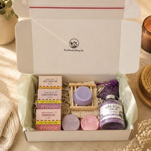Op de afbeelding: Een open doos gevuld met badproducten, waaronder zeep, badzout en een houten zeepbakje. De doos bevat producten met de tekst "Aurora Glow's Body Wash Bar", "Moonlit Waves Conditioner Bar" en "Sunrise Radiance's Shampoo Bar". De merknaam "EcoHavenShop NL" is zichtbaar.