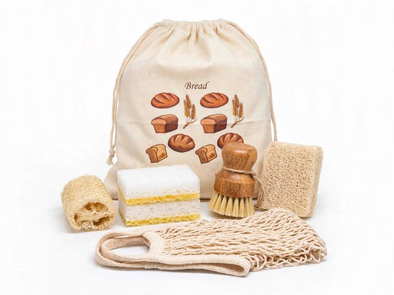 May include: A beige drawstring bag with the word Bread printed above illustrations of various loaves and wheat stalks, accompanied by a white and yellow sponge, a round wooden scrub brush, two rectangular loofah sponges, and a beige mesh bag.