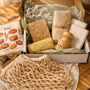 May include: A cardboard box containing a wooden dish brush, two rectangular loofah sponges, a cylindrical loofah, two white square sponges, and a beige cloth. Beside the box is a beige mesh produce bag and a cloth with bread illustrations.