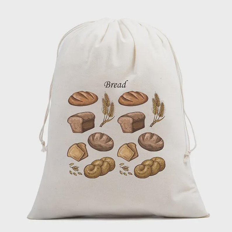 May include: An off-white drawstring bag featuring a printed graphic of various types of bread and wheat stalks. The word Bread is printed in a dark font above the illustrations. The bag is made of a light-colored fabric and has two drawstrings.