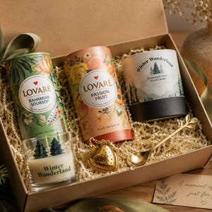 Könnte beinhalten: Eine Geschenkbox mit zwei zylindrischen Teedosen mit der Aufschrift Lovaré Bahamian Soursop und Passion Fruit, einer Winter Wonderland Kerze, einer kleinen Glaskerze mit zwei baumförmigen Wachsen, einem goldenen herzförmigen Tee-Ei und einem goldenen Löffel.