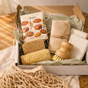 May include: A cardboard box containing a white cloth with bread illustrations and the text Bread, a beige fabric roll tied with twine, two rectangular white sponges, two textured tan loofah sponges, and a wooden dish brush with light bristles.