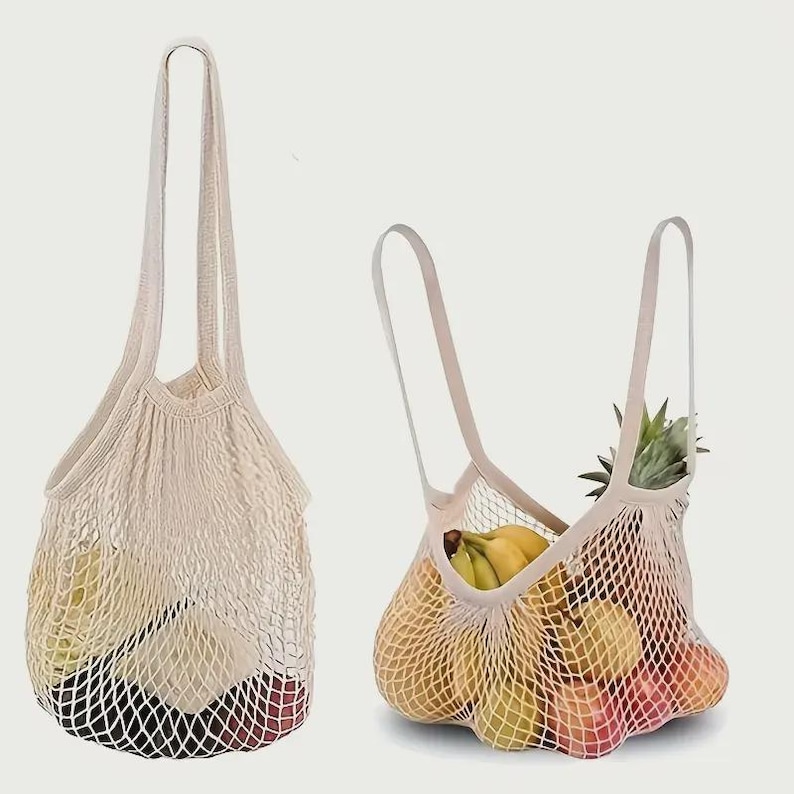 May include: Two off-white reusable mesh bags with long handles. One bag is empty, and the other is filled with various fruits including a pineapple, bananas, apples, and pears, showcasing the expandable net construction of the fabric.