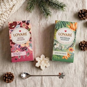 Könnte beinhalten: Zwei rechteckige Schachteln Lovaré-Tee, eine rosa mit Blumen- und Beerenmustern und eine grüne mit Illustrationen von Bahamian Soursop. Eine weiße schneeflockenförmige Kerze, ein silberner Löffel mit Schneeflockengriff und Tannenzapfen liegen bei.