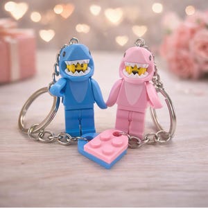 Peut inclure: Deux porte-clés en forme de requin de style Lego, un bleu et un rose, se tenant la main. Chaque figurine a une tête de requin et un visage jaune. Une pièce Lego en forme de cœur relie les porte-clés. L'arrière-plan est flou avec des lumières bokeh.