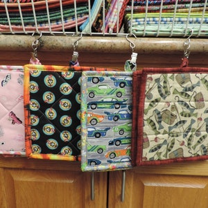 May include: Four colourful quilted pot holders hanging on hooks. The pot holders feature different patterns, including pink with a roller skate design, black with a cartoon character design, grey with a car design, and brown with a fishing design.