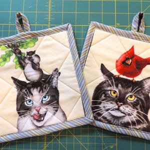 May include: Two quilted pot holders with a cat design. One pot holder features a cat with a squirrel perched on its head. The other pot holder features a cat with a cardinal perched on its head.