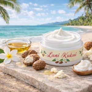 May include: A jar of white cream labeled "Lani Kukui" sits on a stone surface with a wooden spoon, shea nuts, and a bowl of oil. A white flower and green leaves are also present. The background shows a beach and ocean.