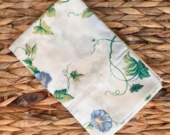 May include: A folded white fabric square featuring a botanical print of blue morning glory flowers, green leaves, and thin curling vines. The textile displays a repeating pattern of floral elements scattered across the light-colored surface.