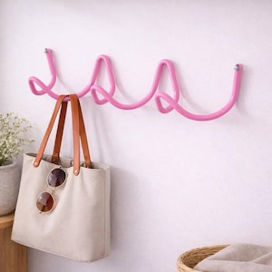 Peut inclure: Un crochet mural en métal rose avec un design ondulé. Un sac fourre-tout beige avec des poignées en cuir marron et des lunettes de soleil est suspendu au crochet. Le mur est peint en blanc.