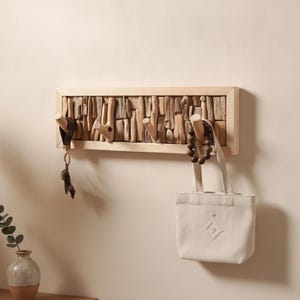 Peut inclure: Un porte-manteau mural rectangulaire doté d'un cadre en bois clair rempli d'une mosaïque de morceaux de bois flotté verticaux. Trois patères en bois lisse et clair dépassent du centre, supportant un porte-clés et un petit sac fourre-tout.
