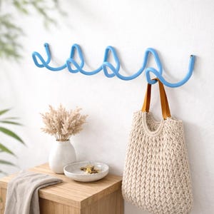 Peut inclure: Un crochet mural en métal bleu clair avec un design ondulé. Un sac fourre-tout tissé beige avec des poignées en cuir marron est suspendu à l'un des crochets. Un vase blanc avec des fleurs séchées et un petit bol sont sur une table en bois.