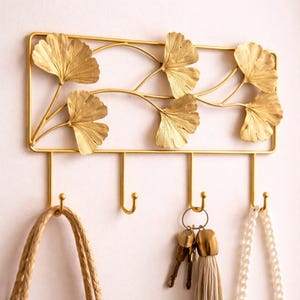 Puede incluir: Un llavero de pared de metal dorado con un marco rectangular y detalles decorativos de hojas de ginkgo. El llavero tiene cuatro ganchos, con una bolsa, llaves y un collar colgando.