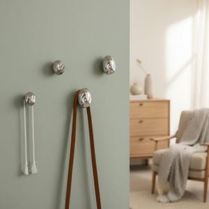 Peut inclure: Quatre crochets muraux argentés au design texturé et organique sont fixés sur un mur vert pâle. L'un des crochets maintient une corde blanche avec des glands, et un autre une sangle en cuir marron. Les crochets ajoutent une touche moderne.