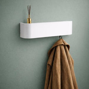 May include: A white wall-mounted shelf with a curved front, holding a gold diffuser bottle with reeds. Below the shelf hangs a brown coat. The shelf is mounted on a muted green wall.