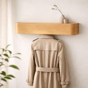 Könnte beinhalten: Helles Holzregal an einer weißen Wand, darunter ein beigefarbener Trenchcoat. Zwei kleine, neutralfarbene Vasen mit einem Zweig Grün stehen auf dem Regal. Das Regal hat eine abgerundete Vorderkante.