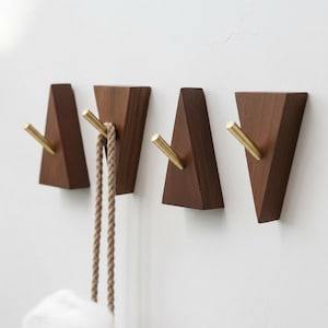May include: Four wooden wall hooks with a triangular design. Each hook is made of dark brown wood and features a gold-colored metal hook. One hook holds a rope, suggesting a storage or organizational theme.