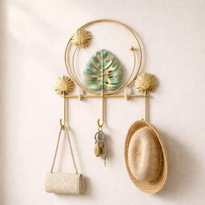 Peut inclure: Un porte-manteau mural en métal doré avec un design circulaire, une feuille turquoise et dorée, trois crochets et des accents floraux décoratifs. Un sac à main beige, des clés et un chapeau de paille sont suspendus aux crochets.