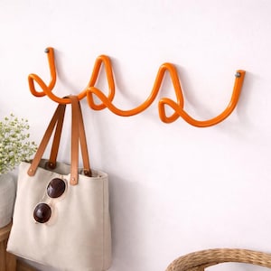 Op de afbeelding: Een oranje metalen wandhaak met een lusontwerp. Een beige draagtas met bruine leren handvatten en zonnebril hangt aan de haak. De haak is aan een witte muur bevestigd.