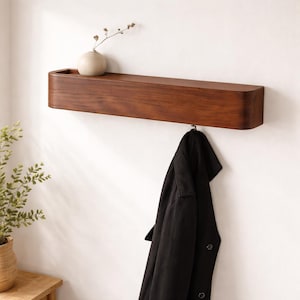 Könnte beinhalten: Dunkelbraunes Wandregal aus Holz mit abgerundeter Vorderkante. Eine kleine, hellfarbene Vase mit einem Zweig getrockneter Blumen steht auf dem Regal. Ein schwarzer Mantel hängt an einem Haken darunter.