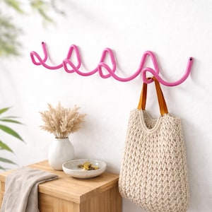 Peut inclure: Un crochet mural en métal rose vif avec cinq boucles. Un sac fourre-tout beige tissé avec des anses en cuir marron est suspendu à l'une des boucles. Le crochet est fixé à un mur blanc, avec une table en bois et des objets décoratifs en arrière-plan.