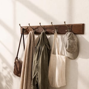 Pode incluir: Um cabide de parede em madeira com cinco ganchos de metal em tom de latão antigo. Pendurados nos ganchos estão uma bolsa de ombro de couro castanho, um cachecol bege, um casaco verde-azeitona, uma sacola de lona creme e um boné cinzento.