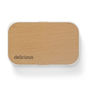 May include: Rectangular lunchbox with a light wood grain top and a white border. The word "delicious" is printed in a small, black font on the lower left corner of the wooden lid. The lunchbox has a simple, modern design.