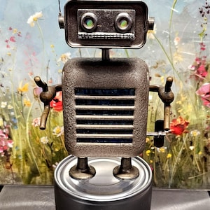 May include: A handcrafted robot sculpture made from repurposed metal components. The robot features a rectangular head with round eyes, a square torso with horizontal lines, and articulated arms. It stands on a cylindrical base, showcasing a unique, industrial aesthetic.
