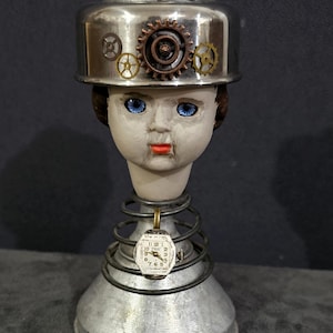 Op de afbeelding: Een steampunk-stijl sculptuur met een poppenhoofd met blauwe ogen en een metalen helm versierd met tandwielen. Een vintage horloge hangt aan een veerbasis, wat een uniek kunstwerk creëert.