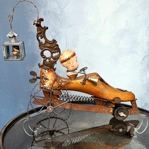 May include: A quirky, handmade sculpture of a wooden shoe repurposed as a vehicle. A doll's head acts as the driver, complete with a small bell and steering wheel. A tiny cage with a doll's head hangs from a curved metal arm. The sculpture has a steampunk style.