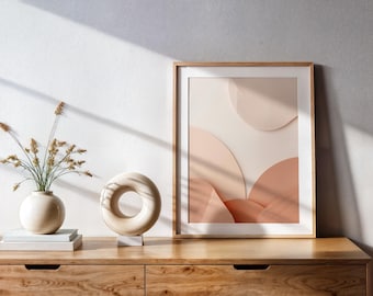 May include: A framed abstract art print with a light wood frame, featuring soft peach and cream-colored shapes. A cream-colored vase with dried flowers and a circular sculpture sit on a wooden cabinet. The scene is lit by natural light.