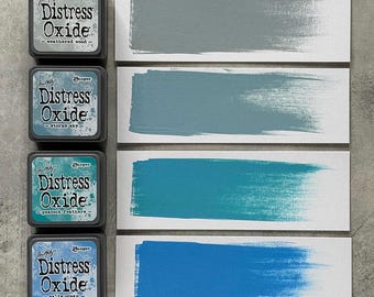 Può includere: Quattro tamponi di inchiostro Distress Oxide con campioni di colore. I tamponi di inchiostro sono etichettati "weathered wood", "stormy sky", "peacock feathers" e "salty ocean". Ogni campione mostra una pennellata del colore corrispondente su carta bianca.